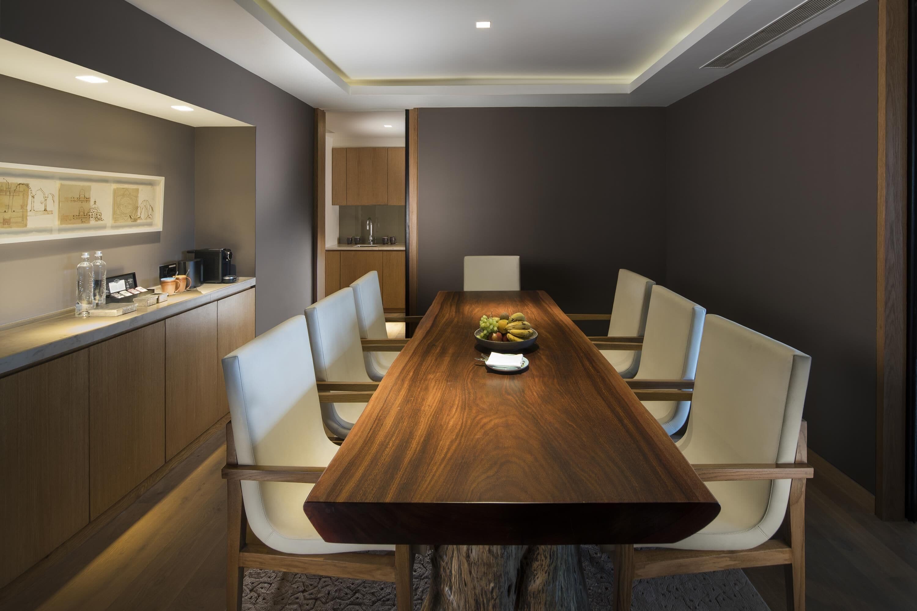 A modern dining area with a long wooden table, white upholstered chairs, and a kitchen area visible in the background.