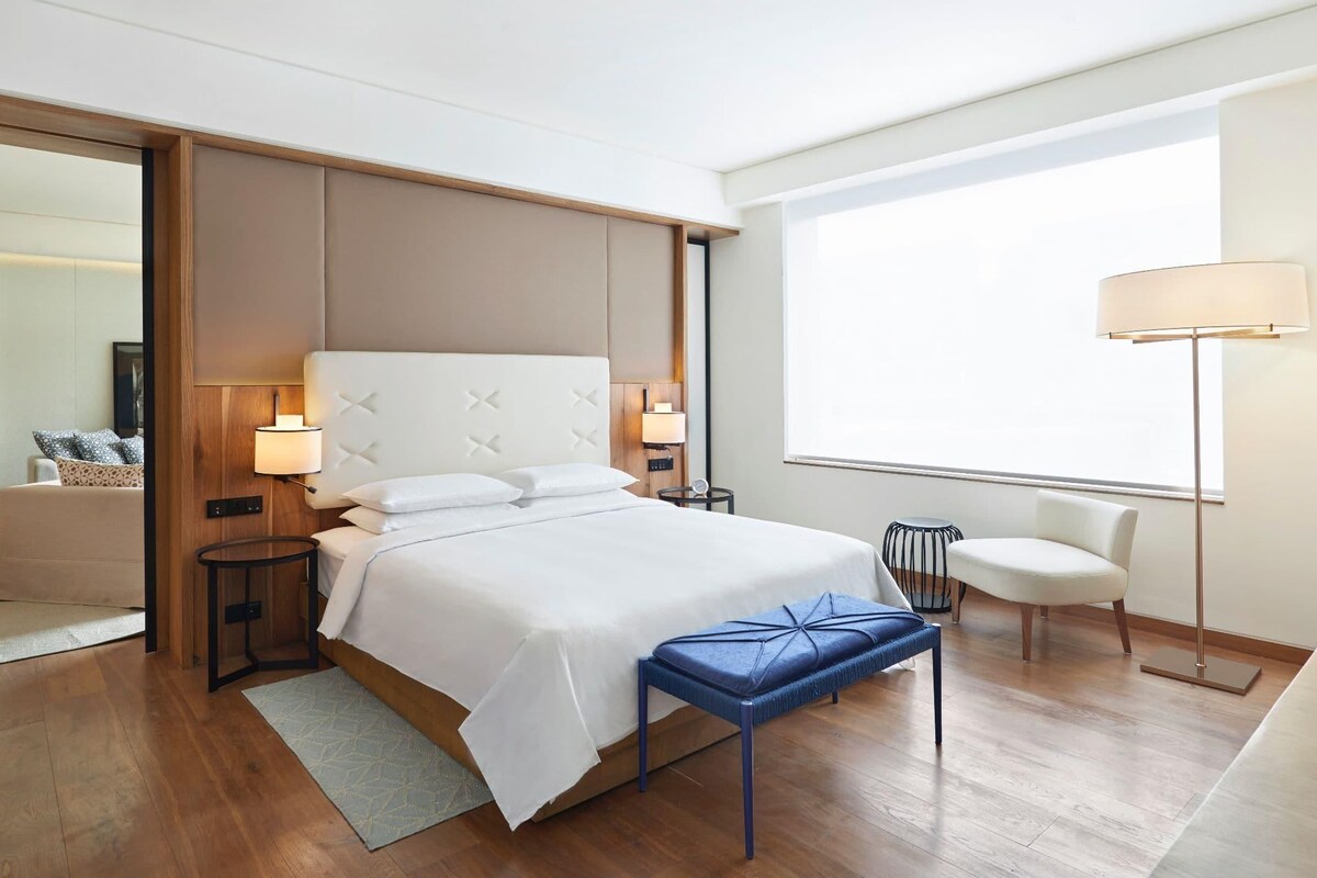 A modern hotel room with a large bed, bedside lamps, a small table, a chair, and a view of the city.