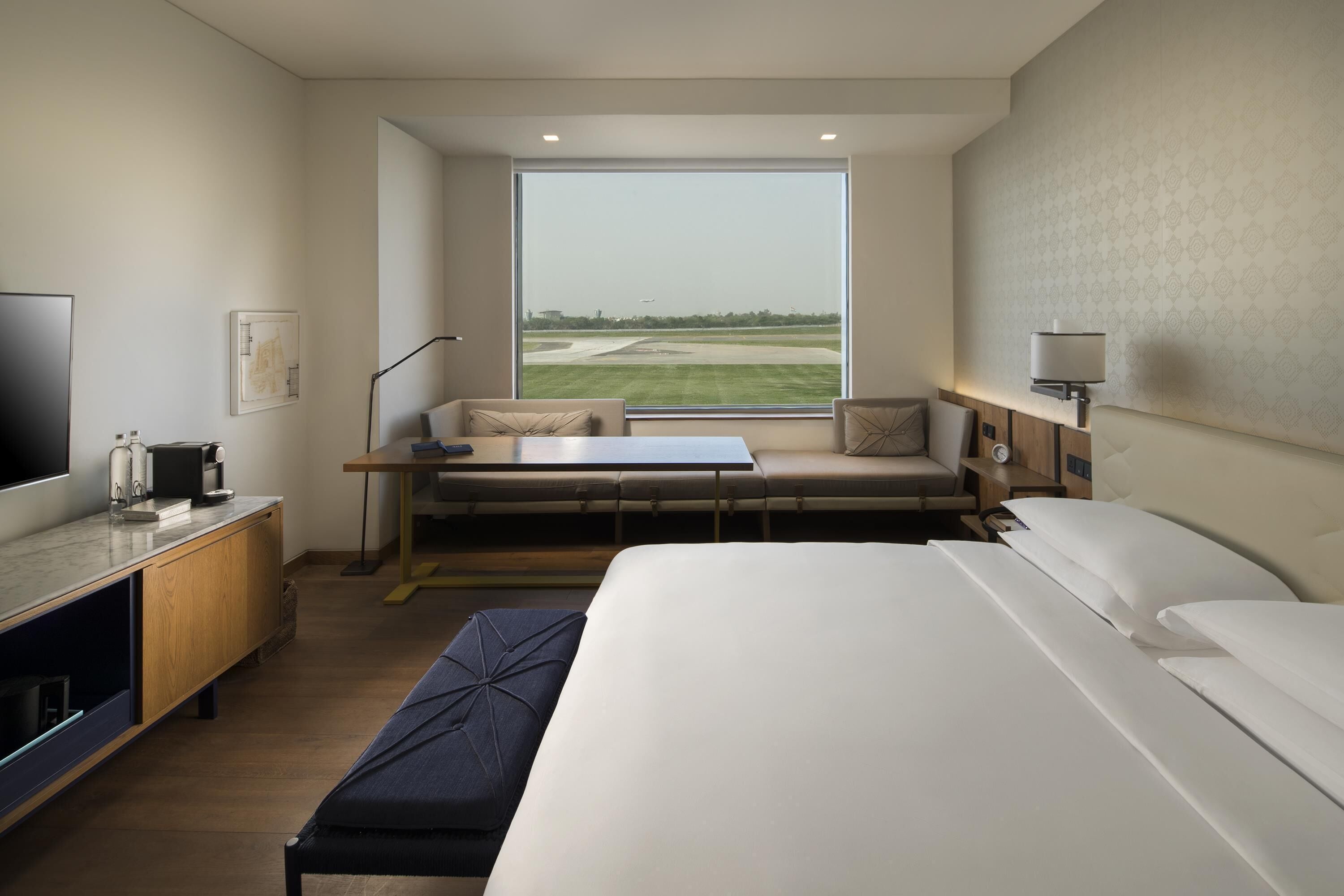 A hotel room with a large bed, a desk with a chair, a sofa, a TV, and a window with a view of a runway.