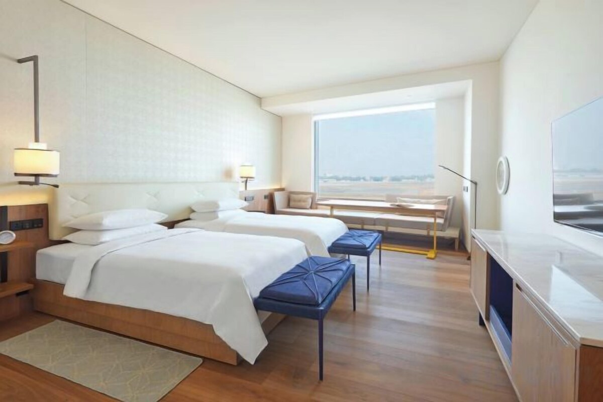 A hotel room with two beds, a desk, and a large window offering a view.