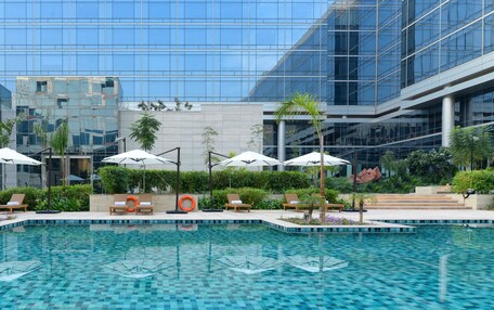 A modern hotel pool area with lounge chairs and umbrellas, surrounded by lush greenery and a glass building facade.