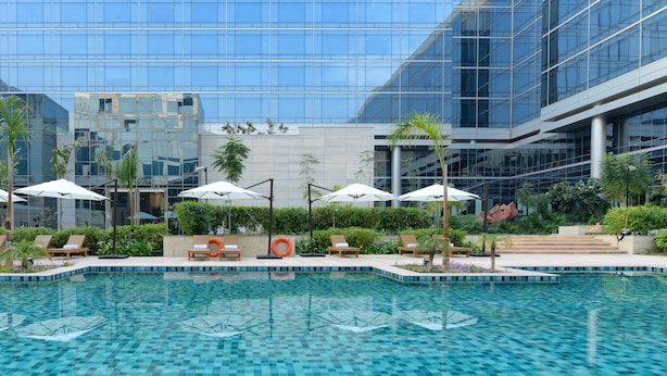 A modern hotel pool area with lounge chairs and umbrellas, surrounded by lush greenery and a glass building facade. - Andaz Delhi, by Hyatt (New Delhi)