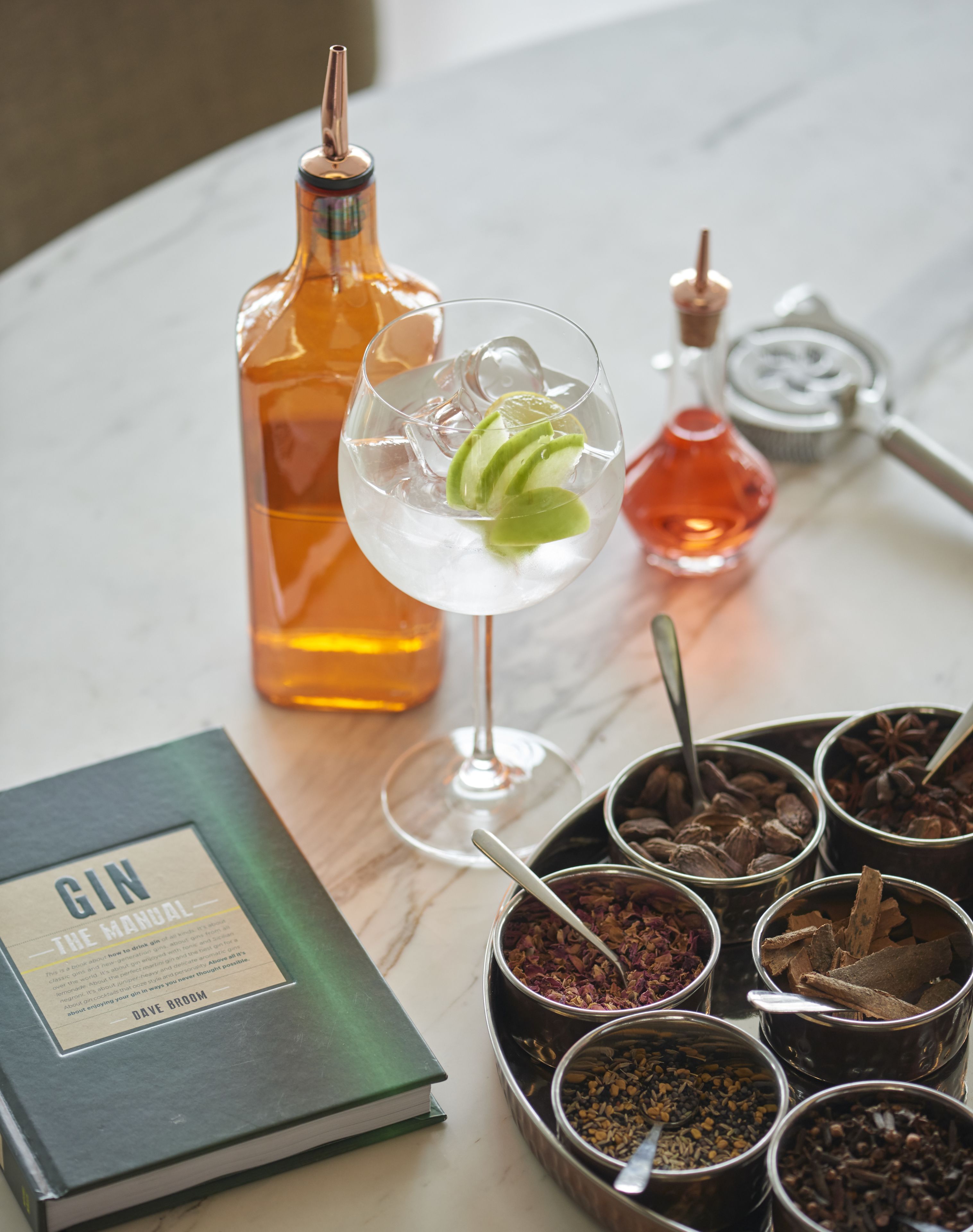 Un tavolo con una bottiglia di gin, un bicchiere di cocktail al lime e diverse spezie in contenitori di metallo.