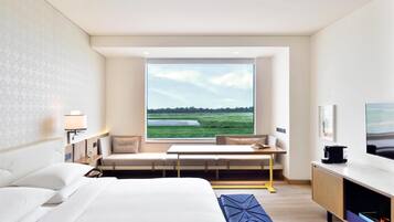 A modern hotel room with a large bed, a seating area with a view of a green landscape, a flat-screen TV, and a coffee machine.