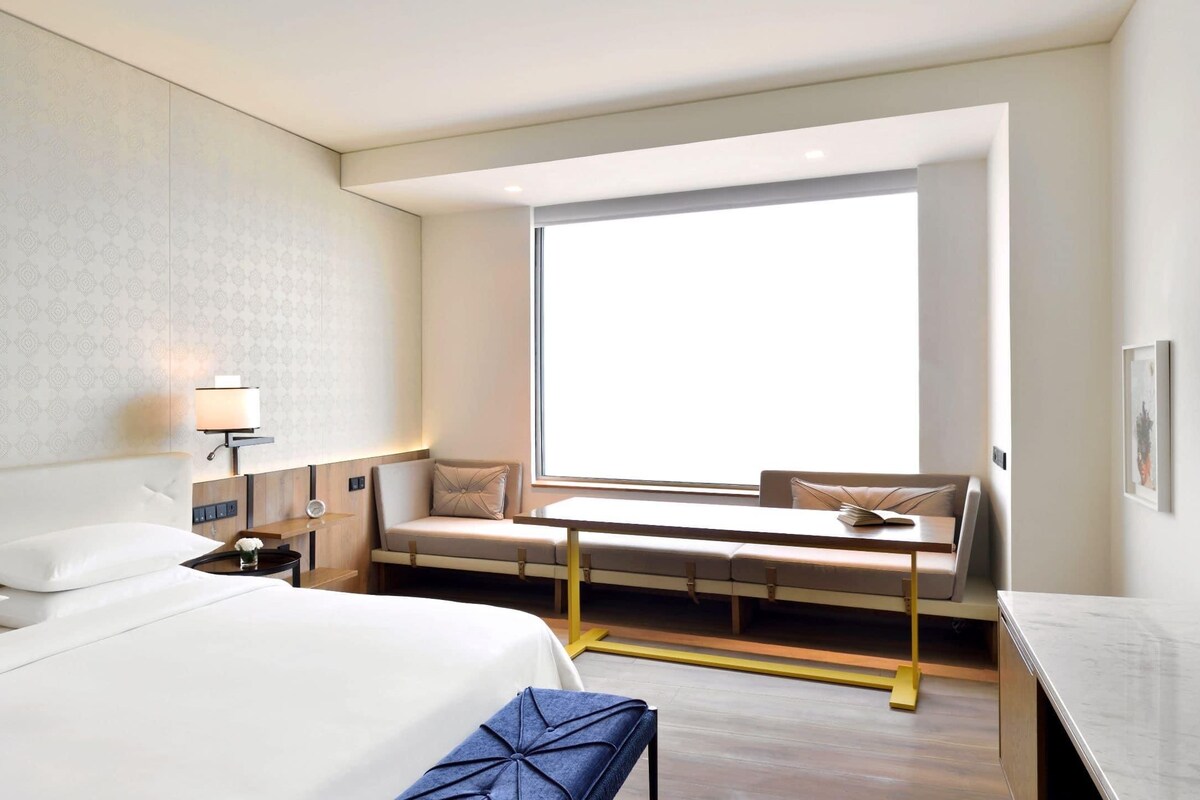 A hotel room with a large bed, a desk with a chair, a window, and a wall-mounted lamp.