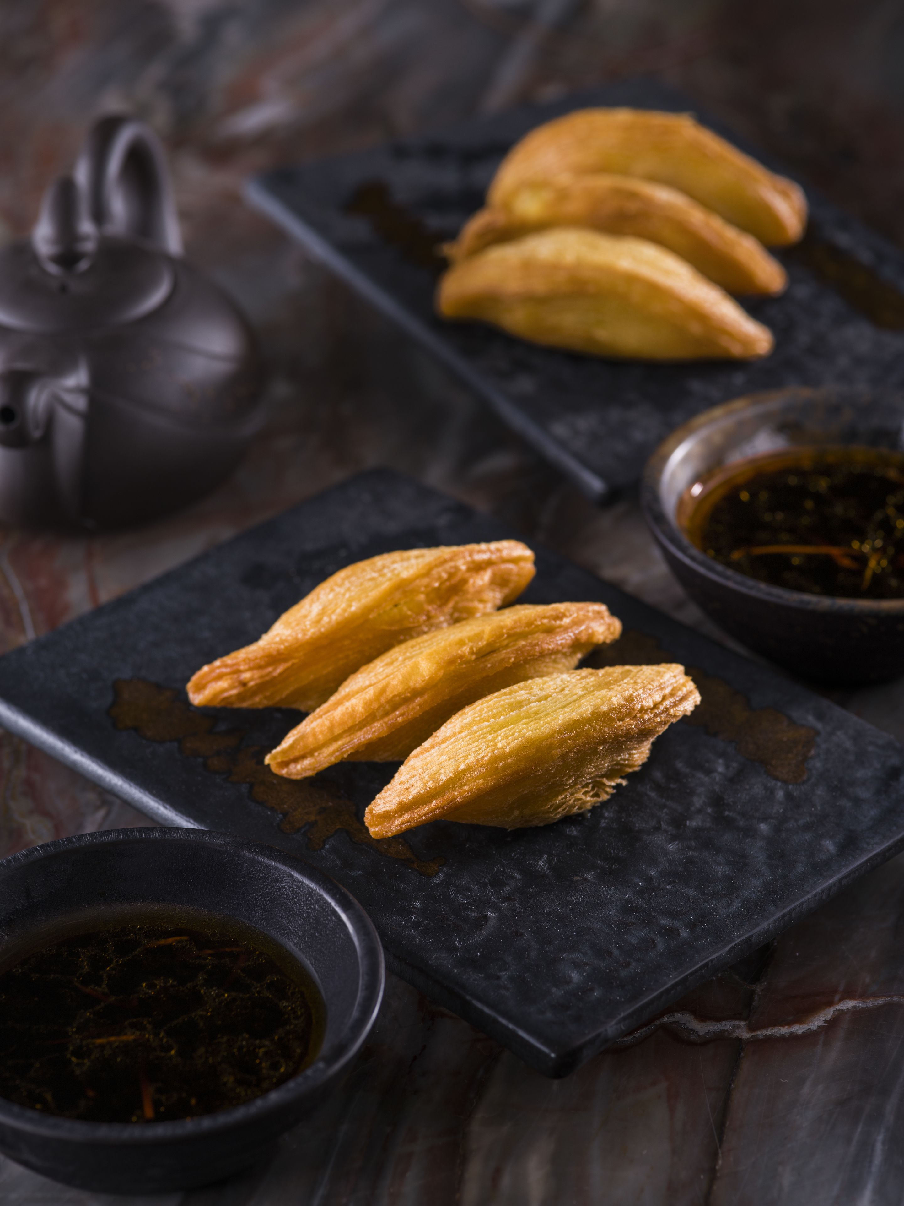 Una bandeja de pizarra oscura con tres pasteles dorados, un recipiente pequeño de salsa oscura y una tetera.