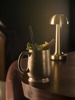 A metallic mug with a spoon and a plant on a wooden surface.