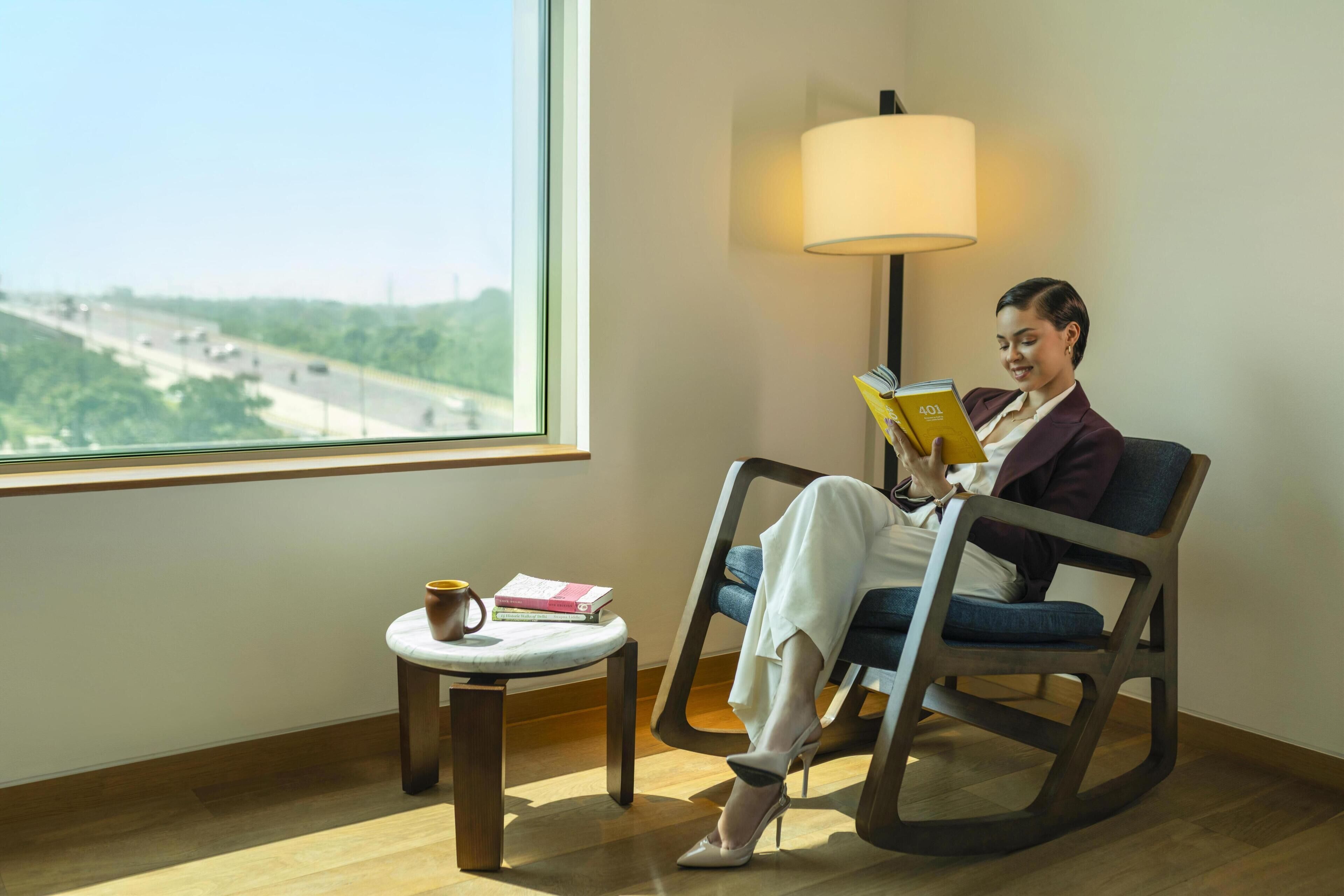 Una persona sentada en una silla leyendo un libro, con una lámpara y una mesita auxiliar con una taza y libros a su lado.