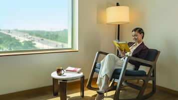 A person seated in a chair reading a book, with a lamp and a small table with a cup and books beside them.