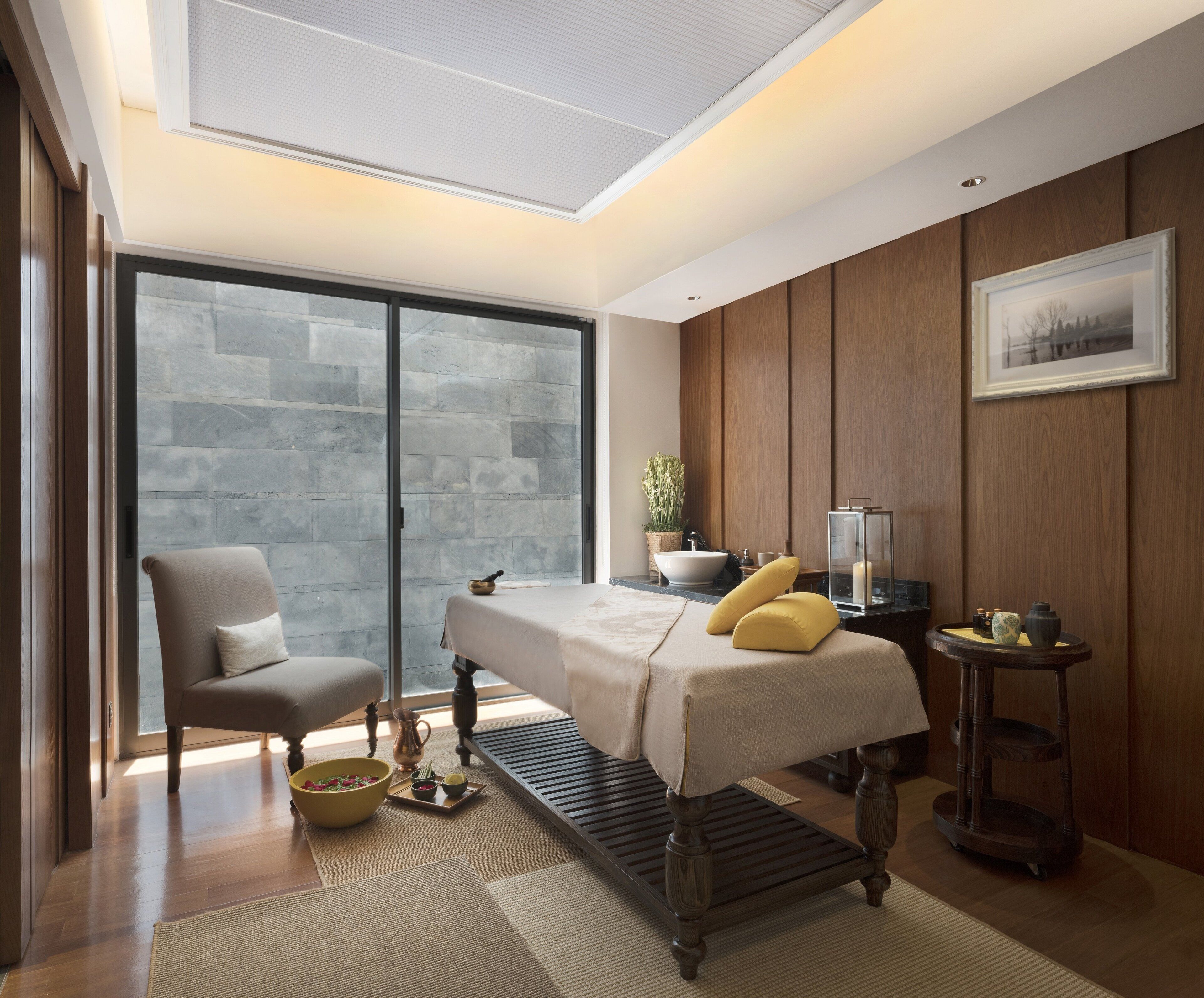 Ein modernes Schlafzimmer mit einer Massageliege, einem Sessel, einem Nachttisch mit Lampe und einem großen Fenster mit Blick auf eine Mauer.