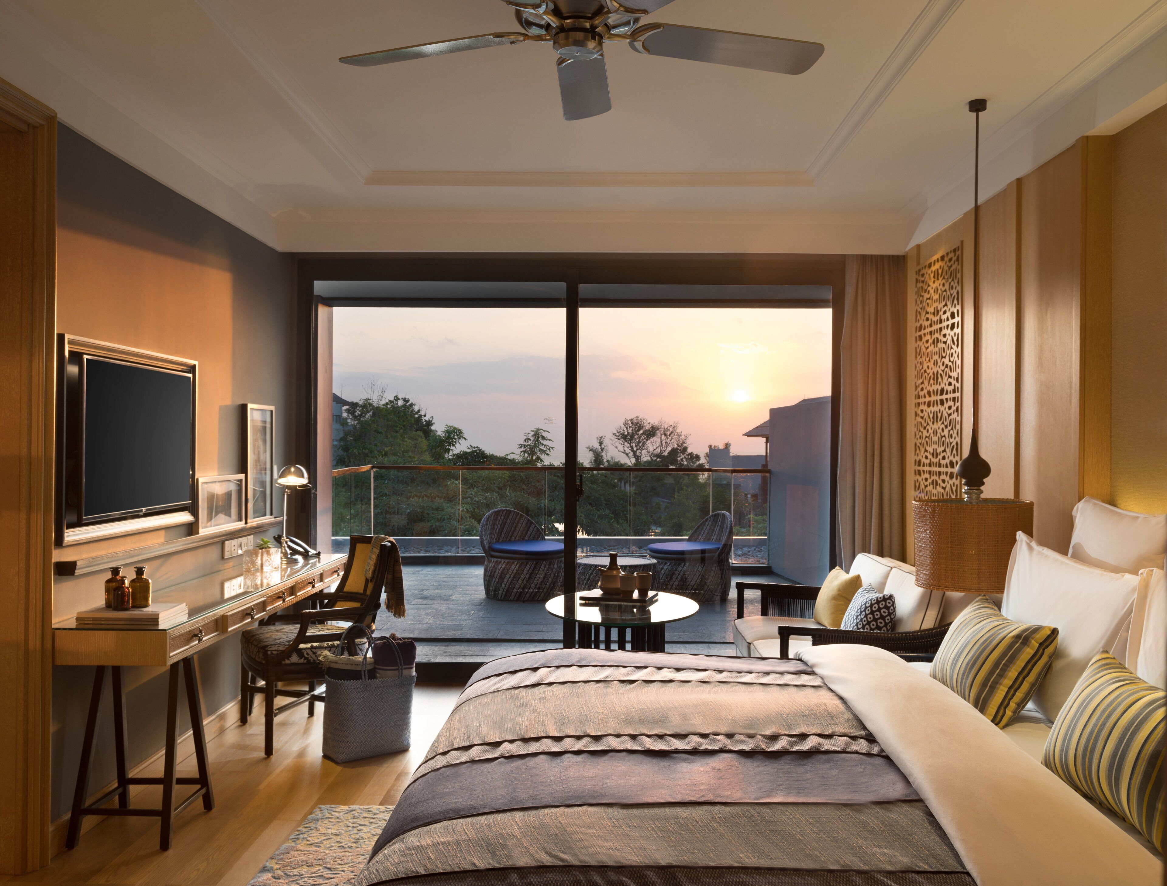 Quarto moderno com cama grande, escrivaninha, cadeira e varanda com vista para o pôr do sol.