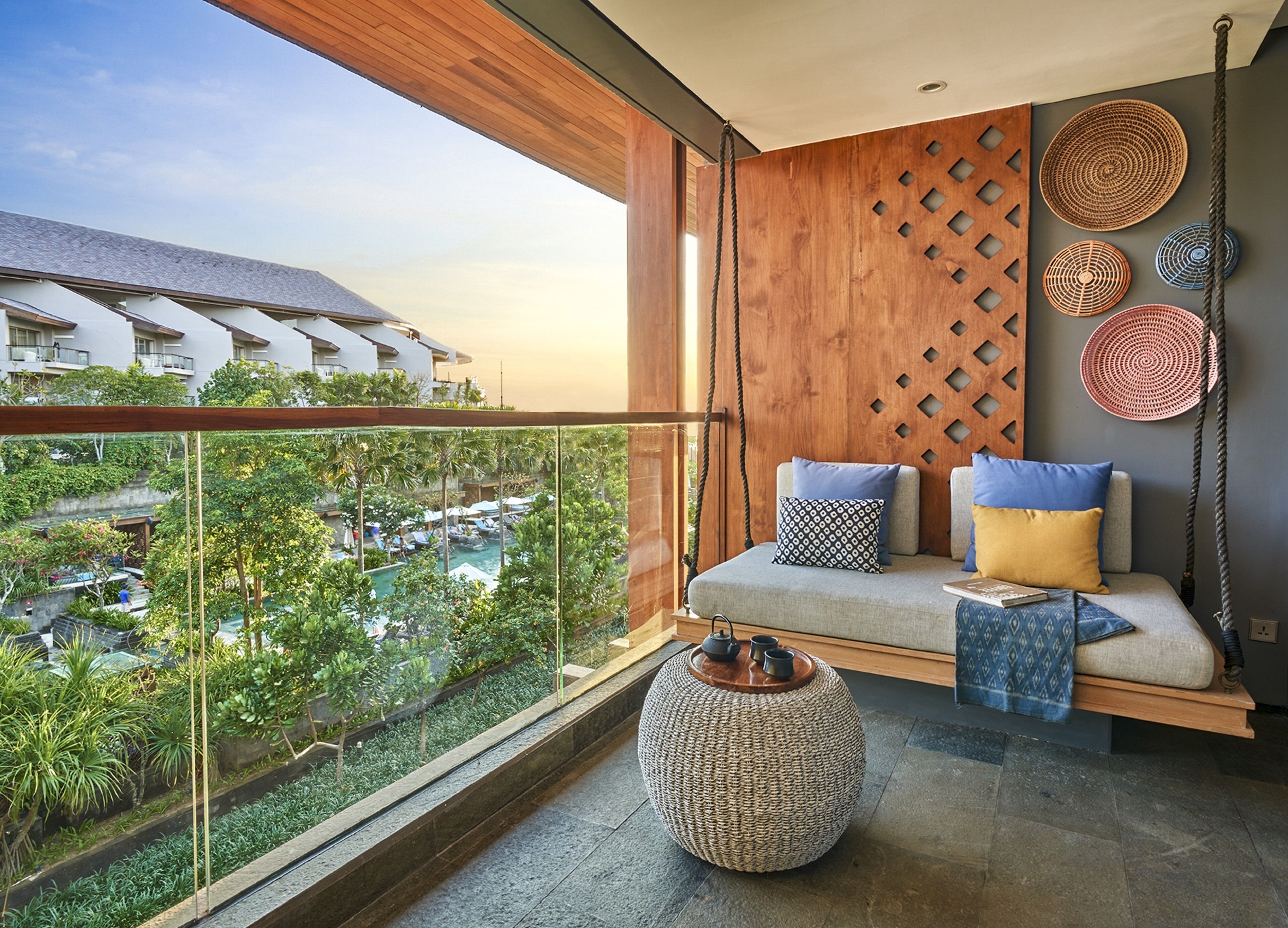 A balcony with a view of a resort, a sofa with cushions, a woven pouf, and decorative wall art.