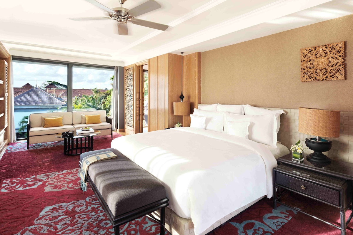 A spacious hotel room with a large bed, a sofa, a bench, a nightstand, and a view of a pool and greenery outside.