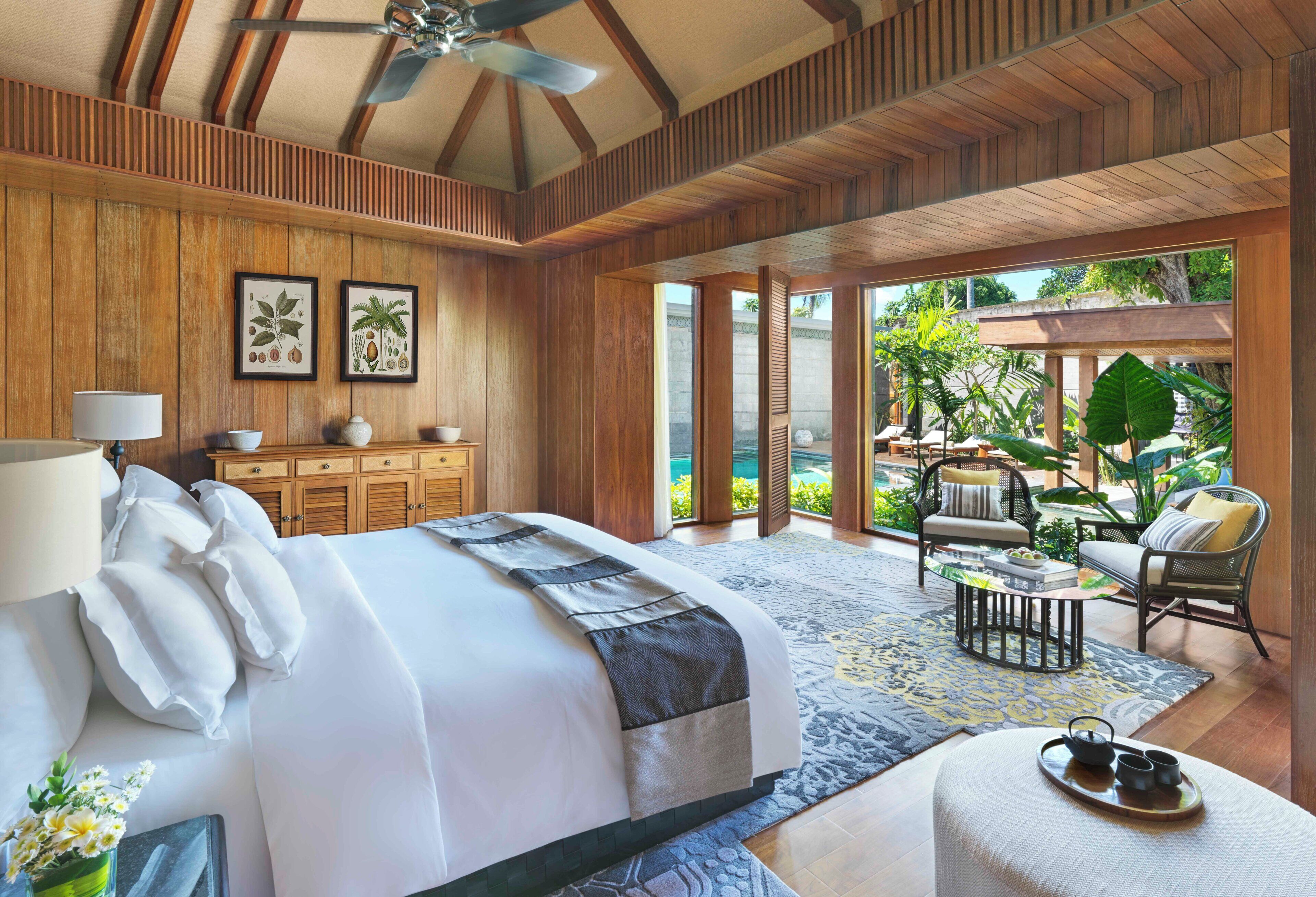 A spacious bedroom with a large bed, a ceiling fan, wooden paneling, and a view of a tropical garden.