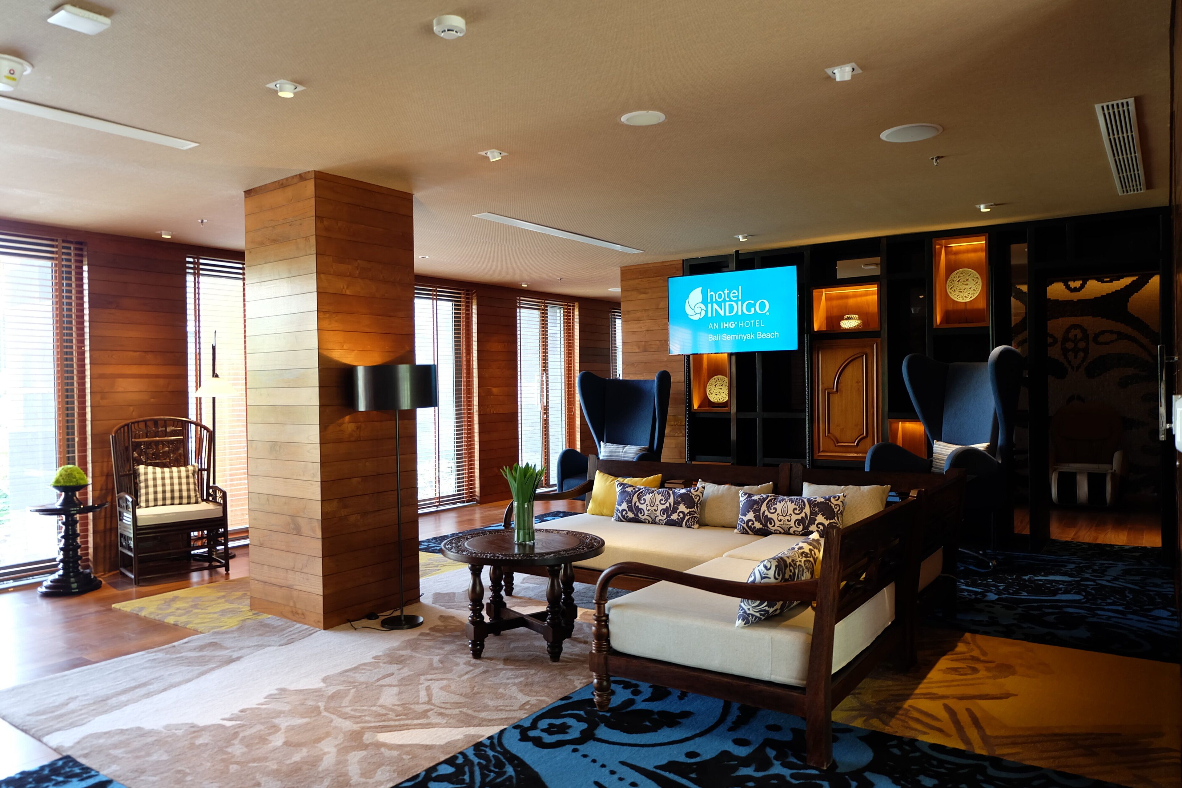 A hotel lobby with a sofa, chairs, a TV screen, and a wooden reception desk.