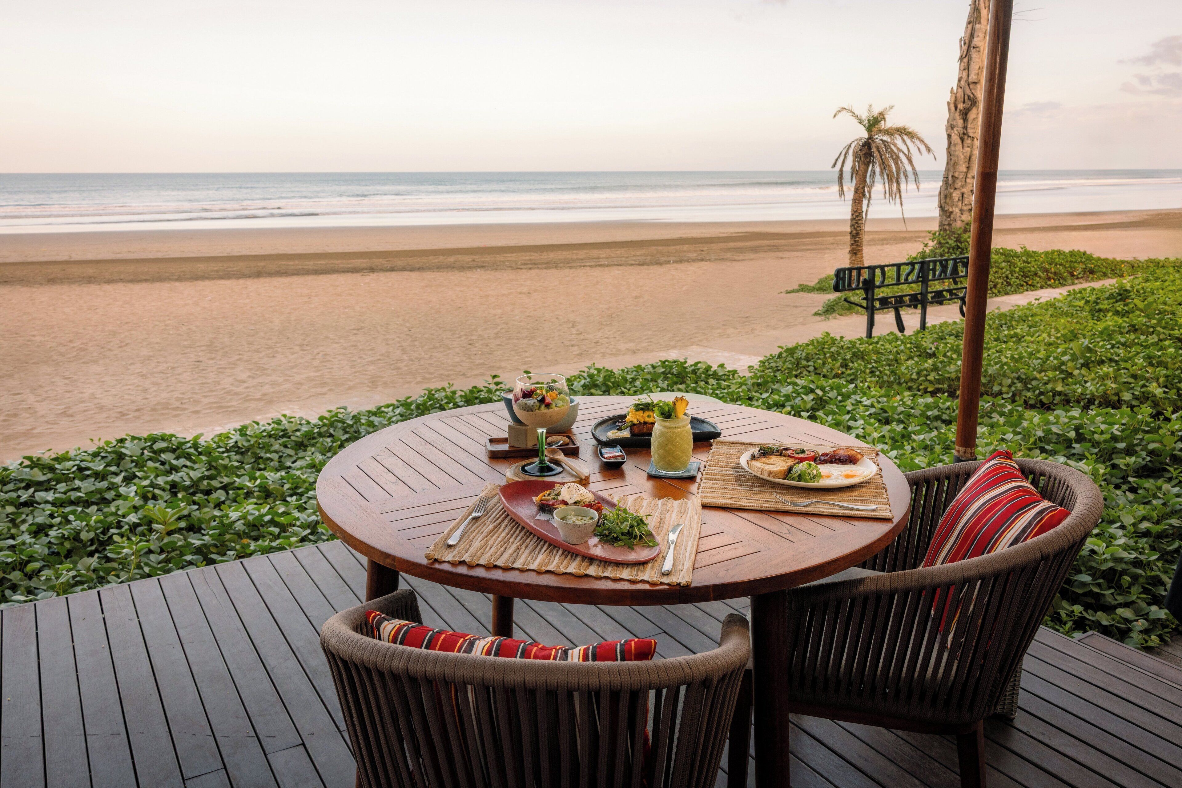 A round wooden table with a woven placemat, surrounded by wicker chairs with striped cushions. The table is set with plates, cutlery, and a glass pitcher. A palm tree and a bench are visible in the background, with a beach and ocean in the distance.