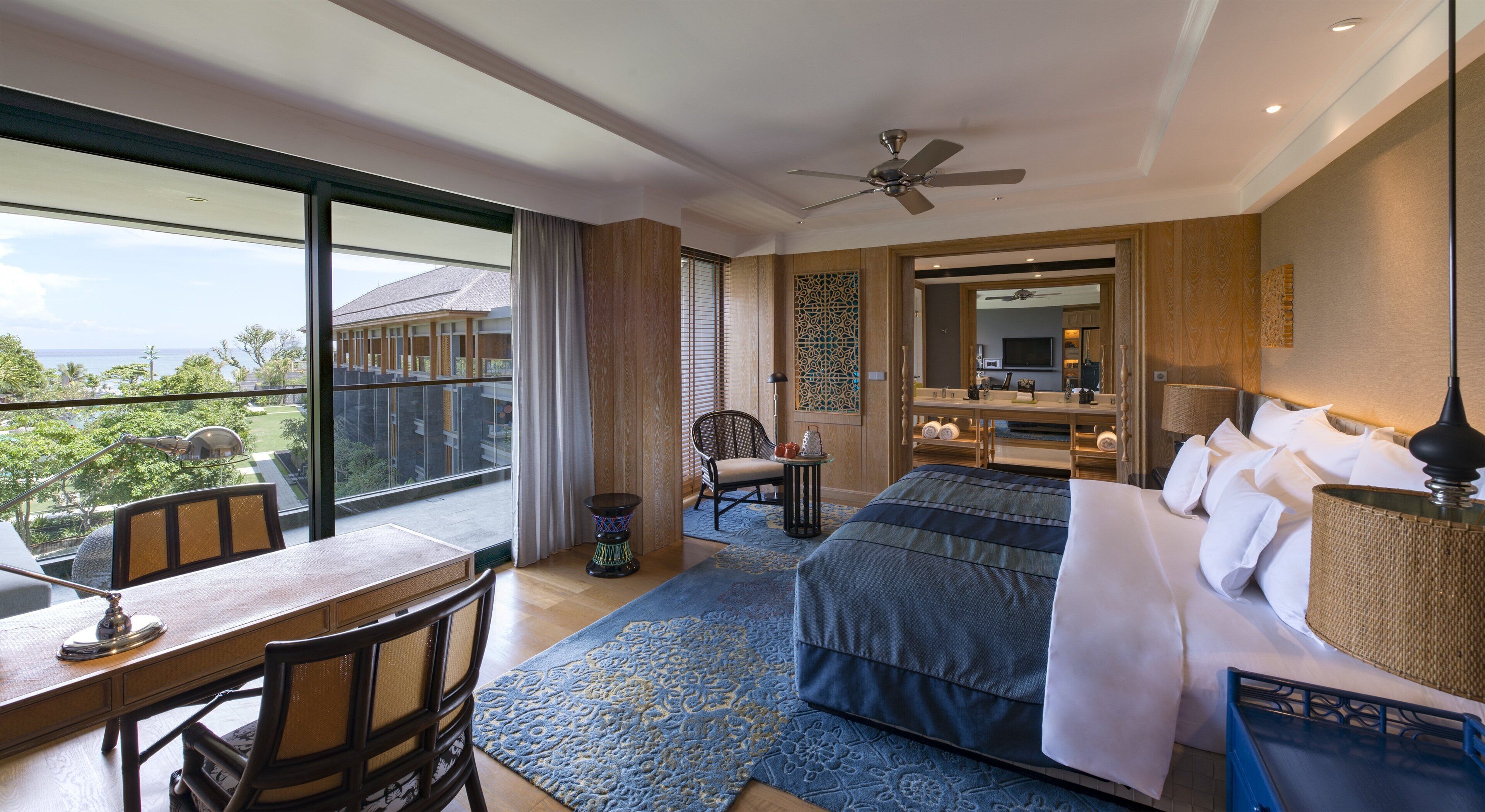 A hotel room with a large balcony, a bed with white linens, a desk with a lamp, and a chair.