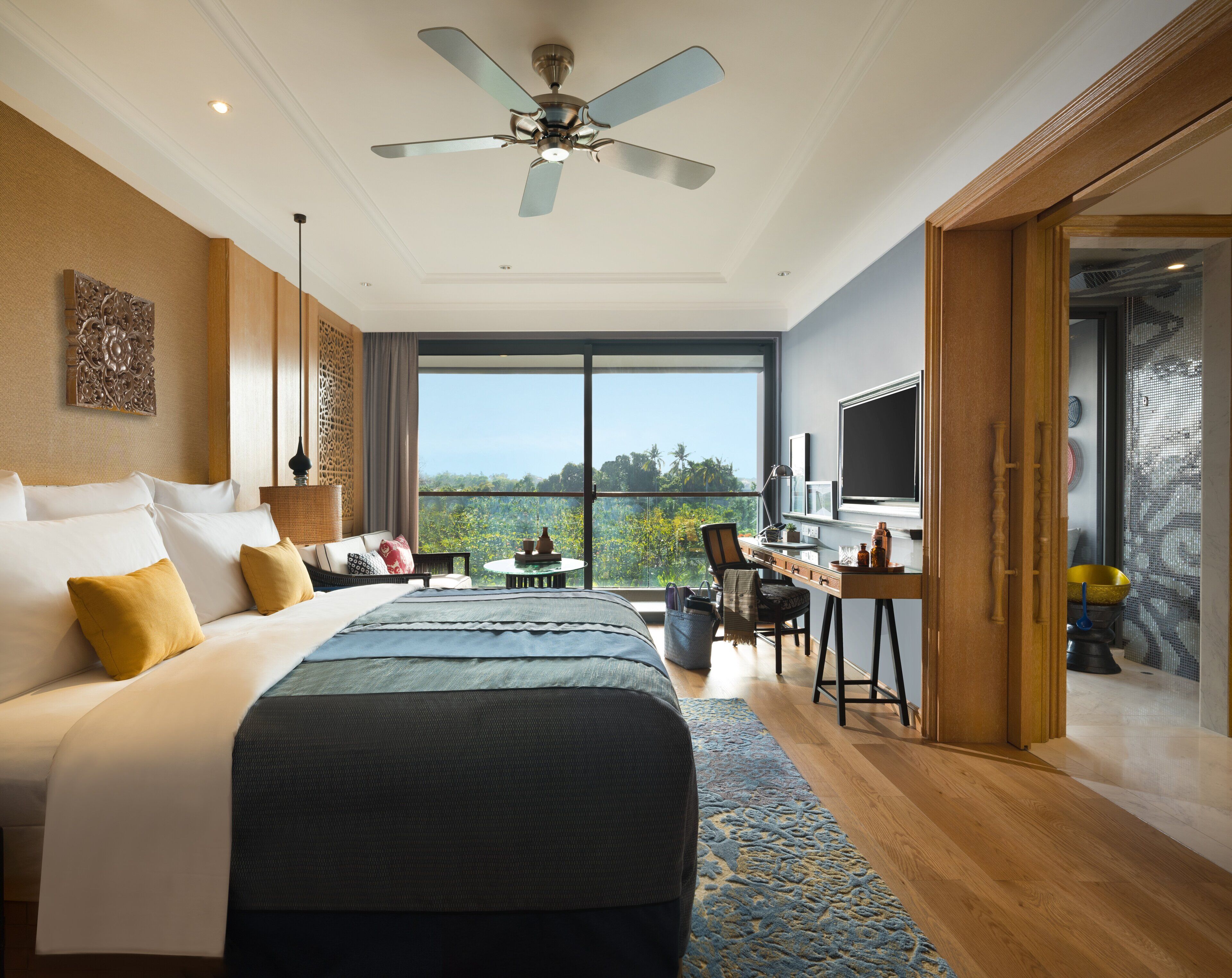 A hotel room with a large bed, a desk with a computer, a television, and a balcony with a view of the trees.