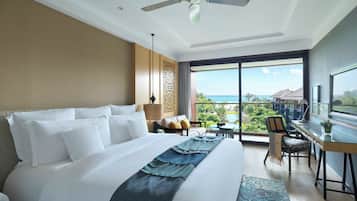 A hotel room with a large bed, a desk, a chair, a view of the ocean, and a ceiling fan.