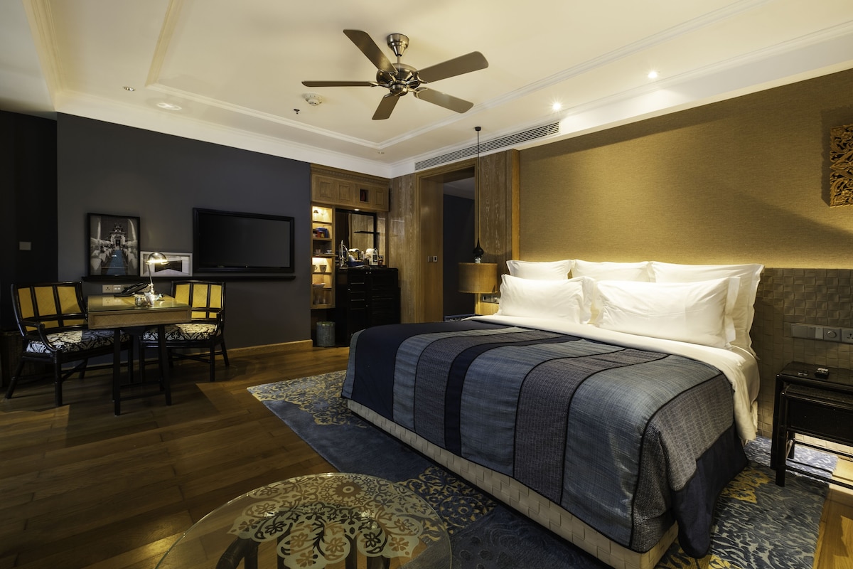 A hotel room with a large bed, a ceiling fan, a TV, a dining area with chairs, and a small table.