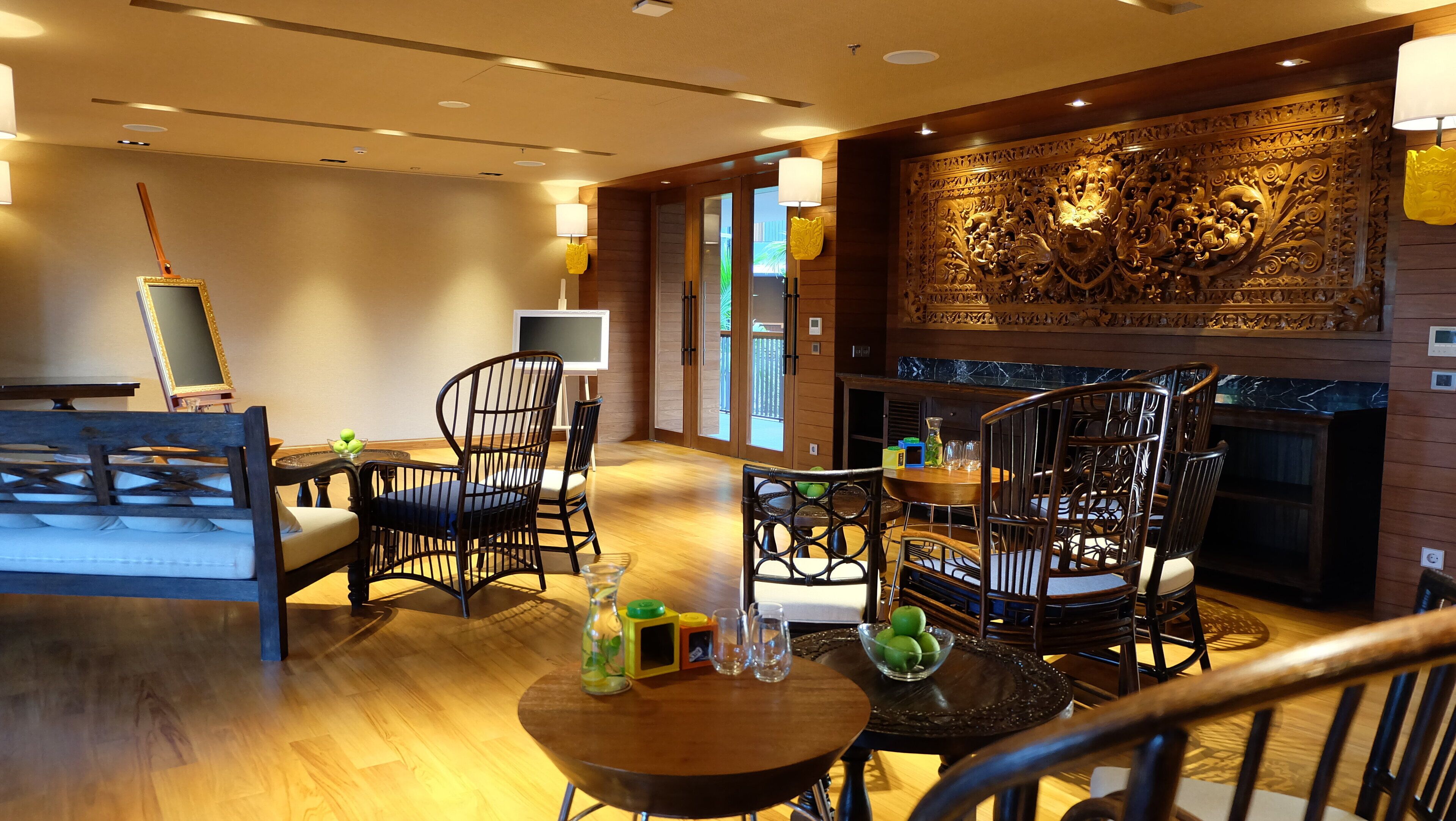A modern lounge area with wooden flooring, black wicker chairs, a round table with a bowl of fruit, and a decorative wall panel.