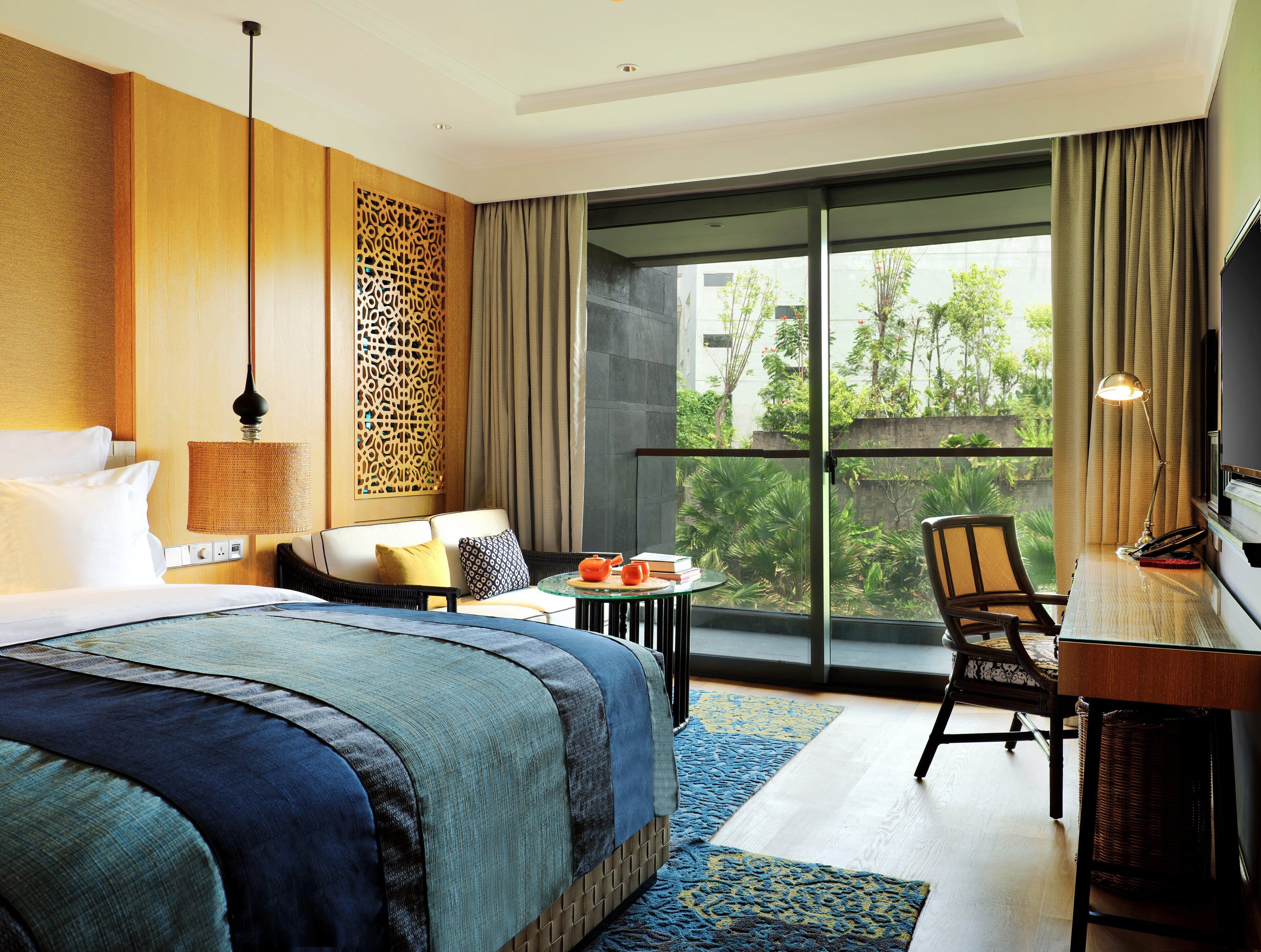 A hotel room with a large bed, a desk with a chair, a table with a teapot, and a view of greenery outside.