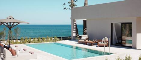 A modern outdoor pool area with a view of the sea, a lounge with sofas and a table, and a covered seating area.