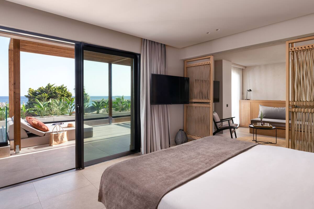 Une chambre d’hôtel moderne dotée d’un grand lit, d’une télévision, d’un canapé, d’un fauteuil et offrant une vue sur l’océan.