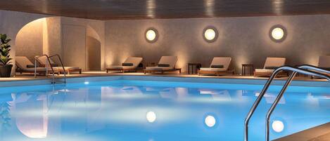 A modern indoor swimming pool with clear water, surrounded by lounge chairs and a metal ladder.