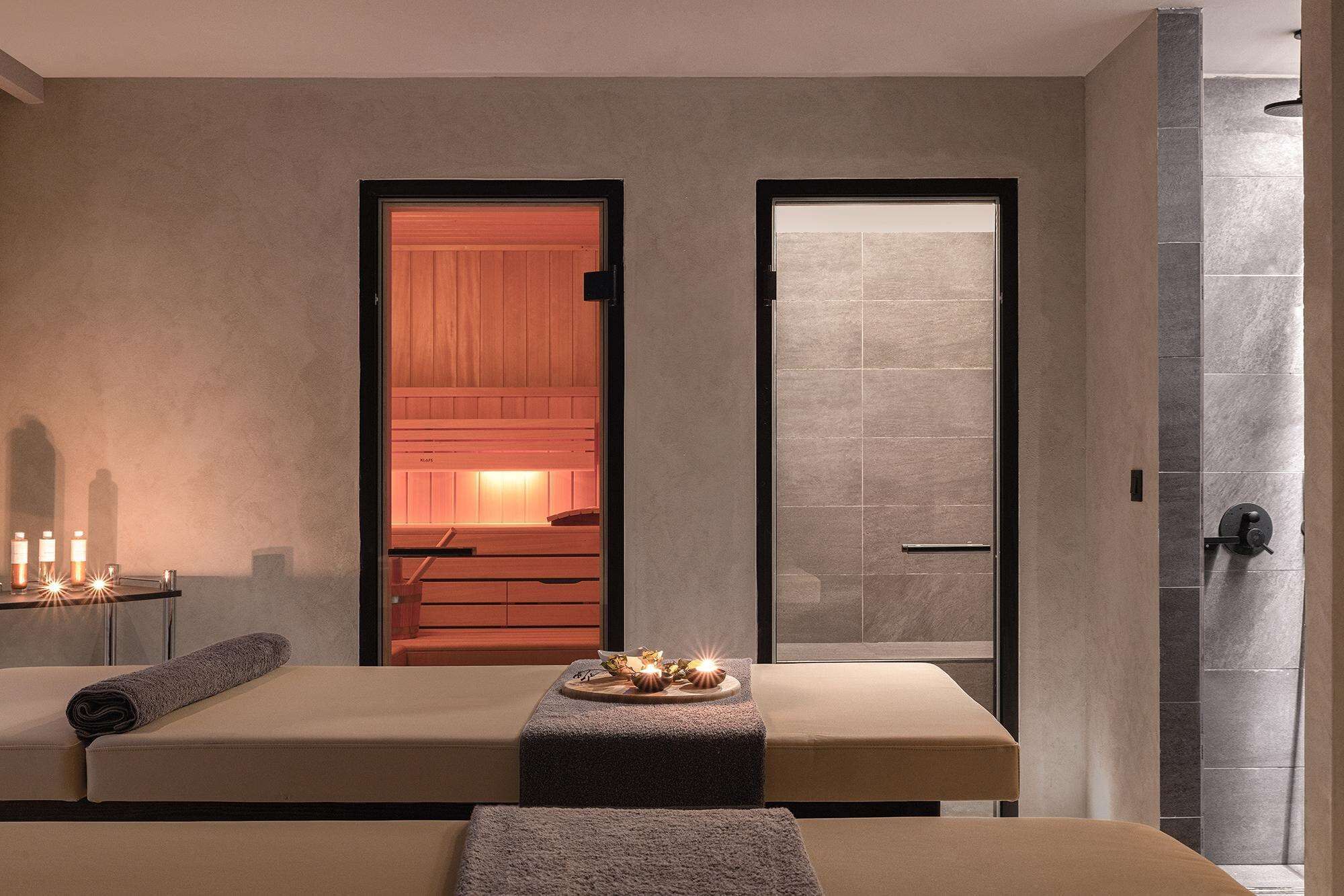 Une salle de spa équipée de deux tables de massage, d’une porte en bois donnant accès à un sauna et d’un espace de douche carrelé.