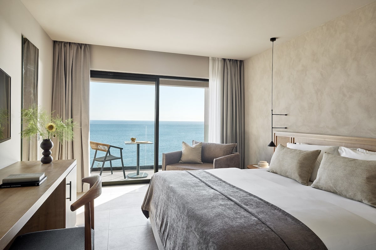 Une chambre d’hôtel avec un grand lit, un bureau et une chaise. La fenêtre offre une vue sur l’océan.