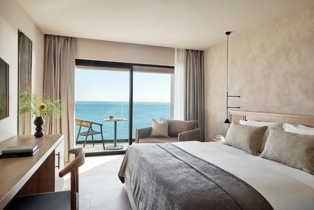 A hotel room with a large bed, a desk, a chair, and a view of the sea.