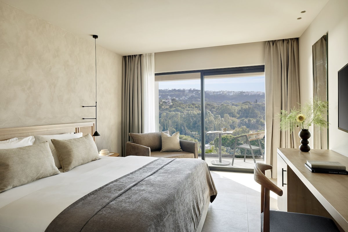 Une chambre d’hôtel moderne, dotée d’un grand lit, d’un bureau et offrant une vue sur le paysage urbain grâce à de grandes fenêtres.