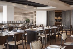 A modern dining area with multiple tables and chairs, a patterned wall, and a visible kitchen area in the background.