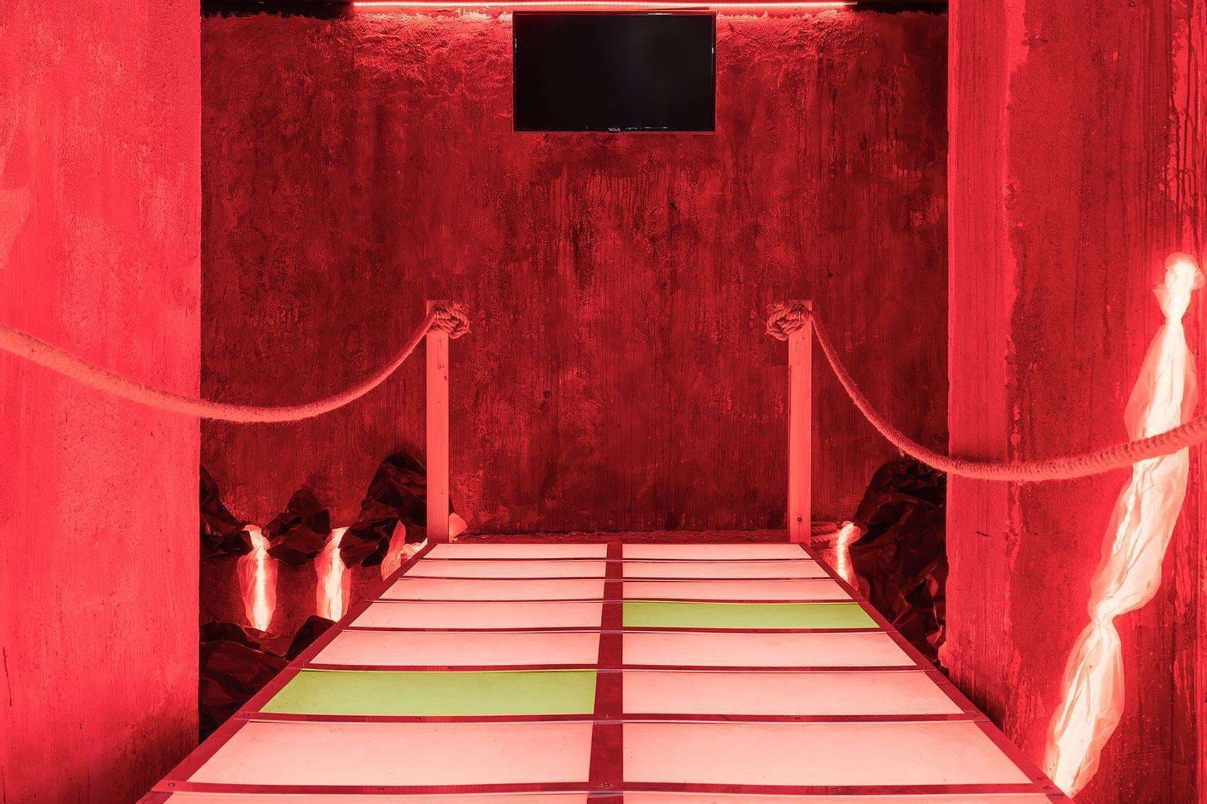 A red-walled room with a bed, a flat-screen TV, and a rope barrier.
