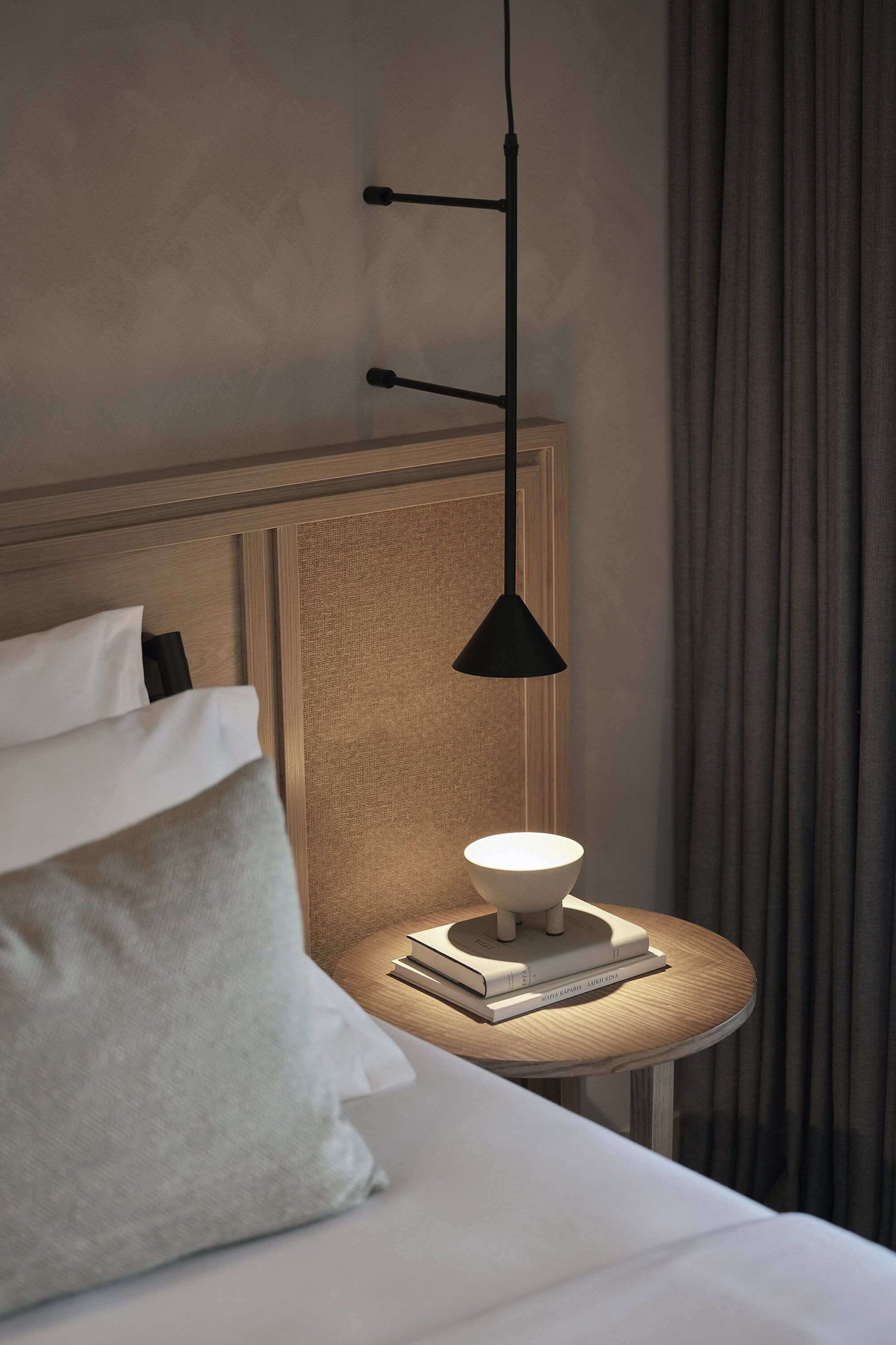Una cama bien hecha con sábanas y almohadones blancos, una mesita de noche con lámpara y libros, y vistas a una cortina y una pared.