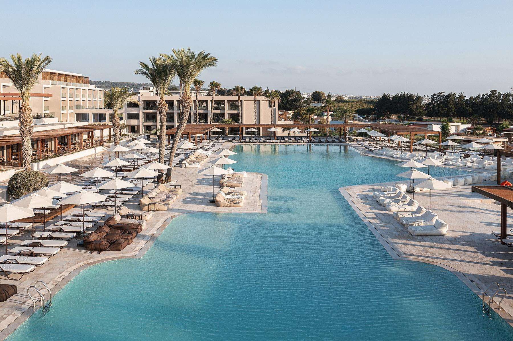 Un área de piscina en un resort con múltiples sillas de descanso y sombrillas, rodeada de palmeras y edificios modernos.