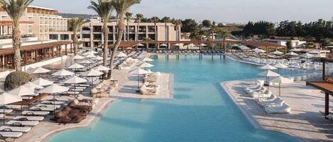 Un área de piscina en un resort con múltiples sillas de descanso y sombrillas, rodeada de palmeras y edificios modernos.