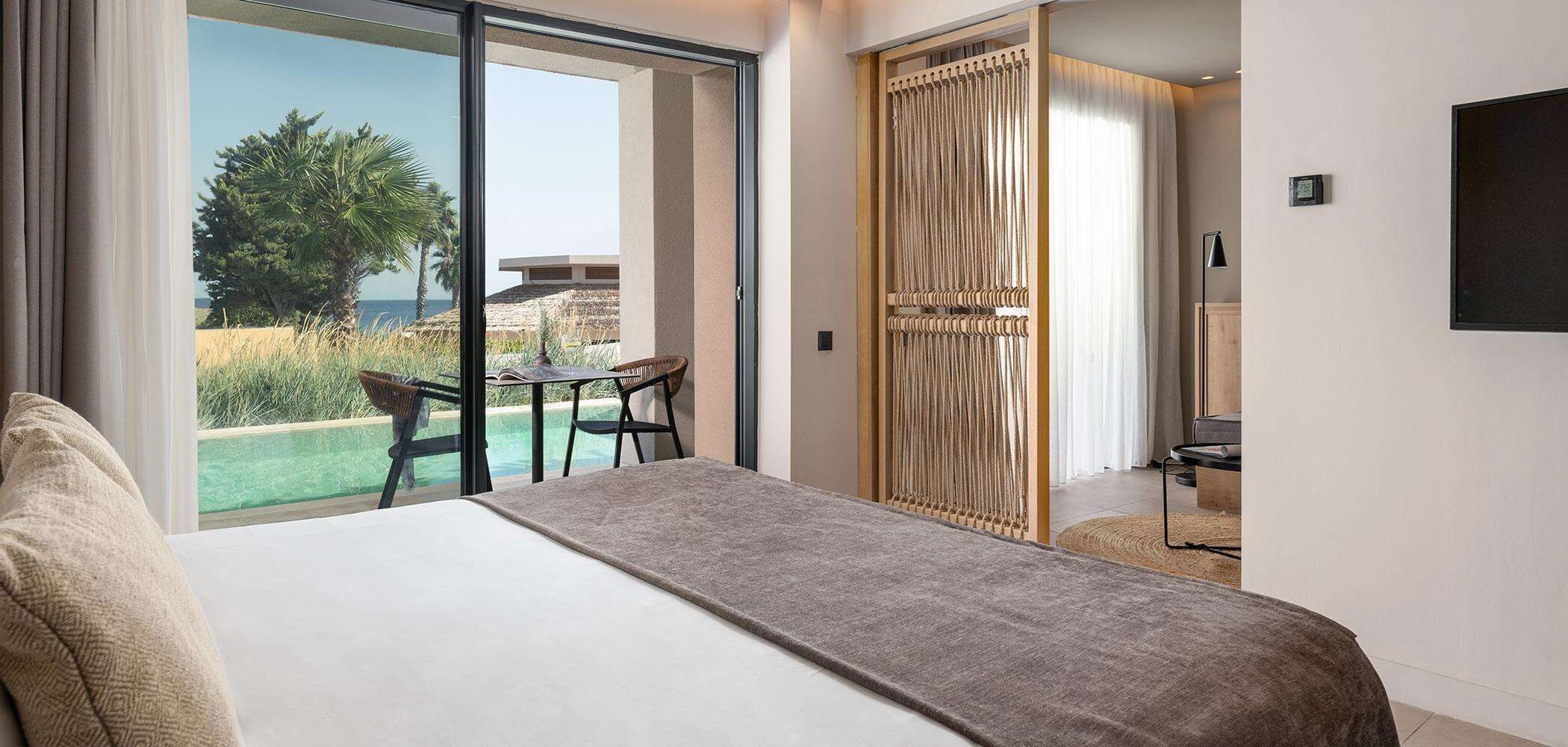 Ein Hotelzimmer mit Bett, kleinem Tisch und Stuhl. Eine Glastür führt zu einem Balkon mit Blick aufs Meer und Palmen.