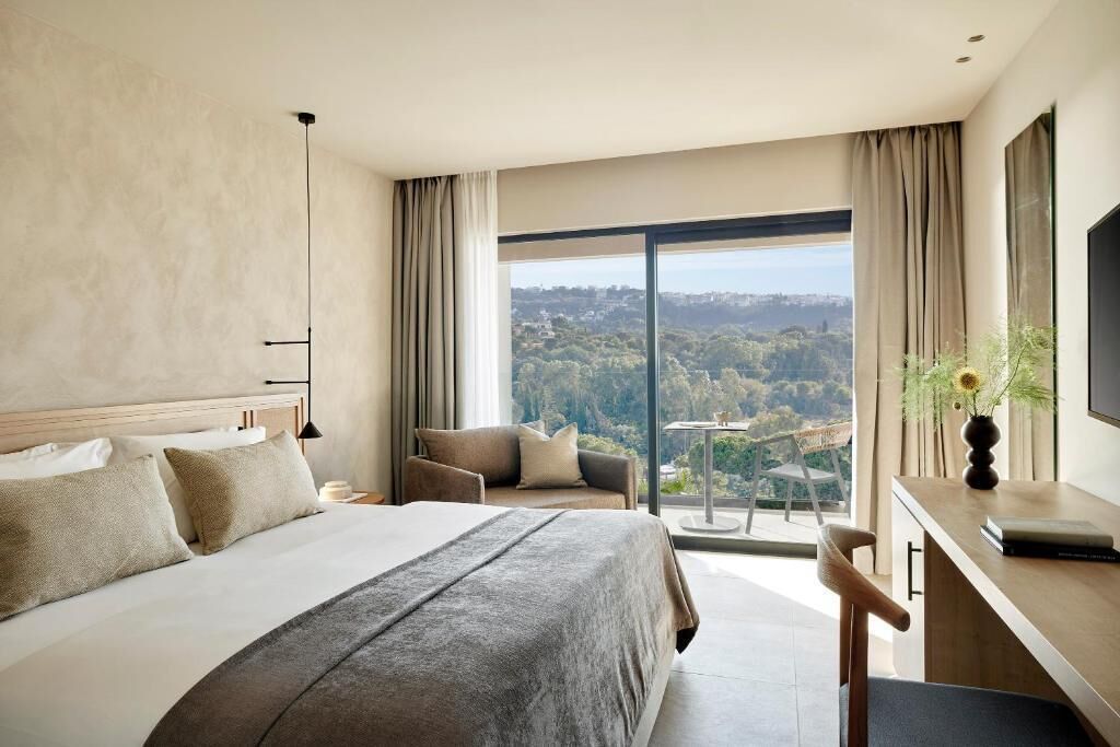A hotel room with a large bed, a desk, a chair, and a balcony with a city view.