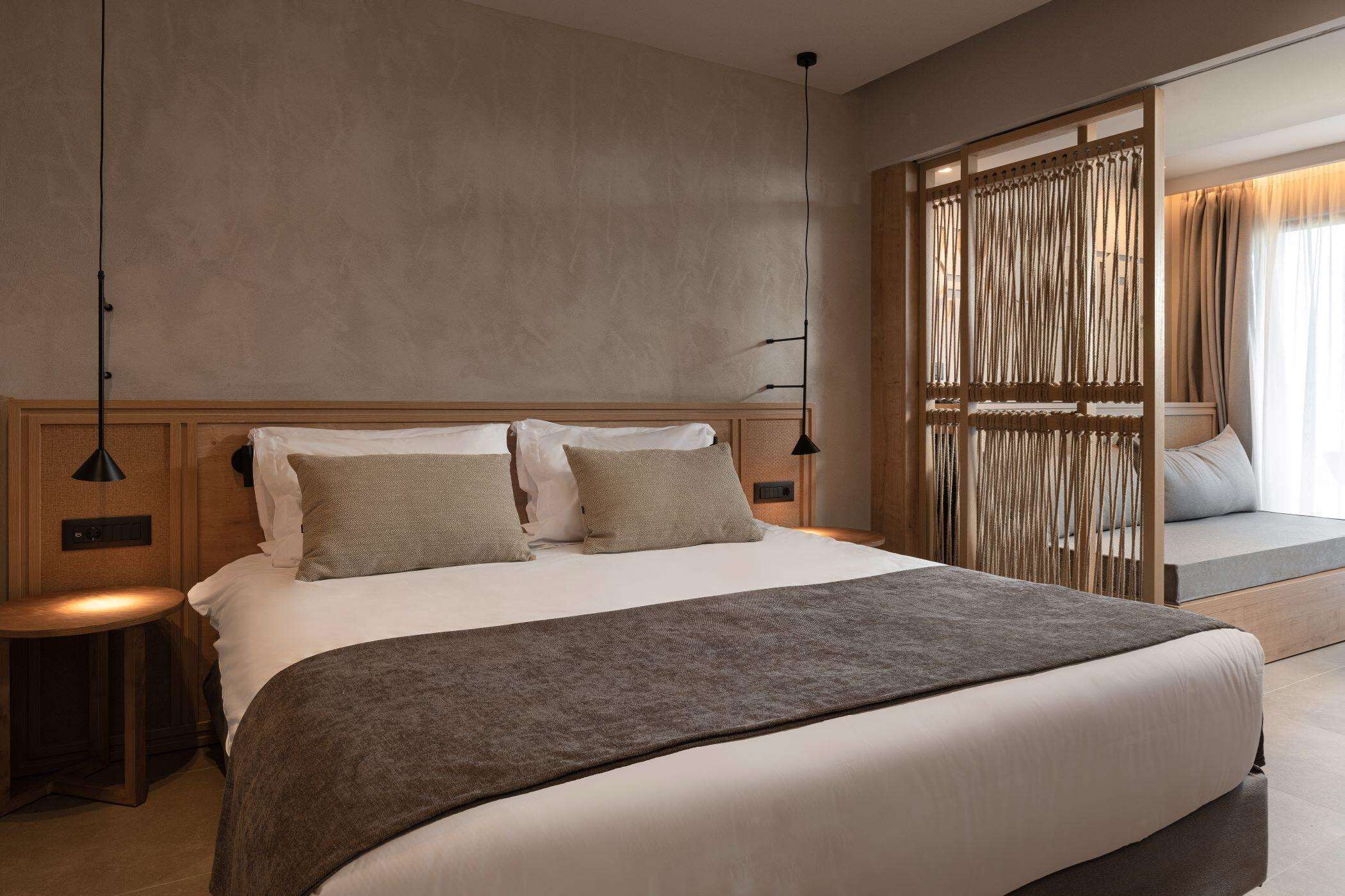 A modern hotel room with a large bed, a bedside table, a lamp, and a view of the outside through a window with curtains.