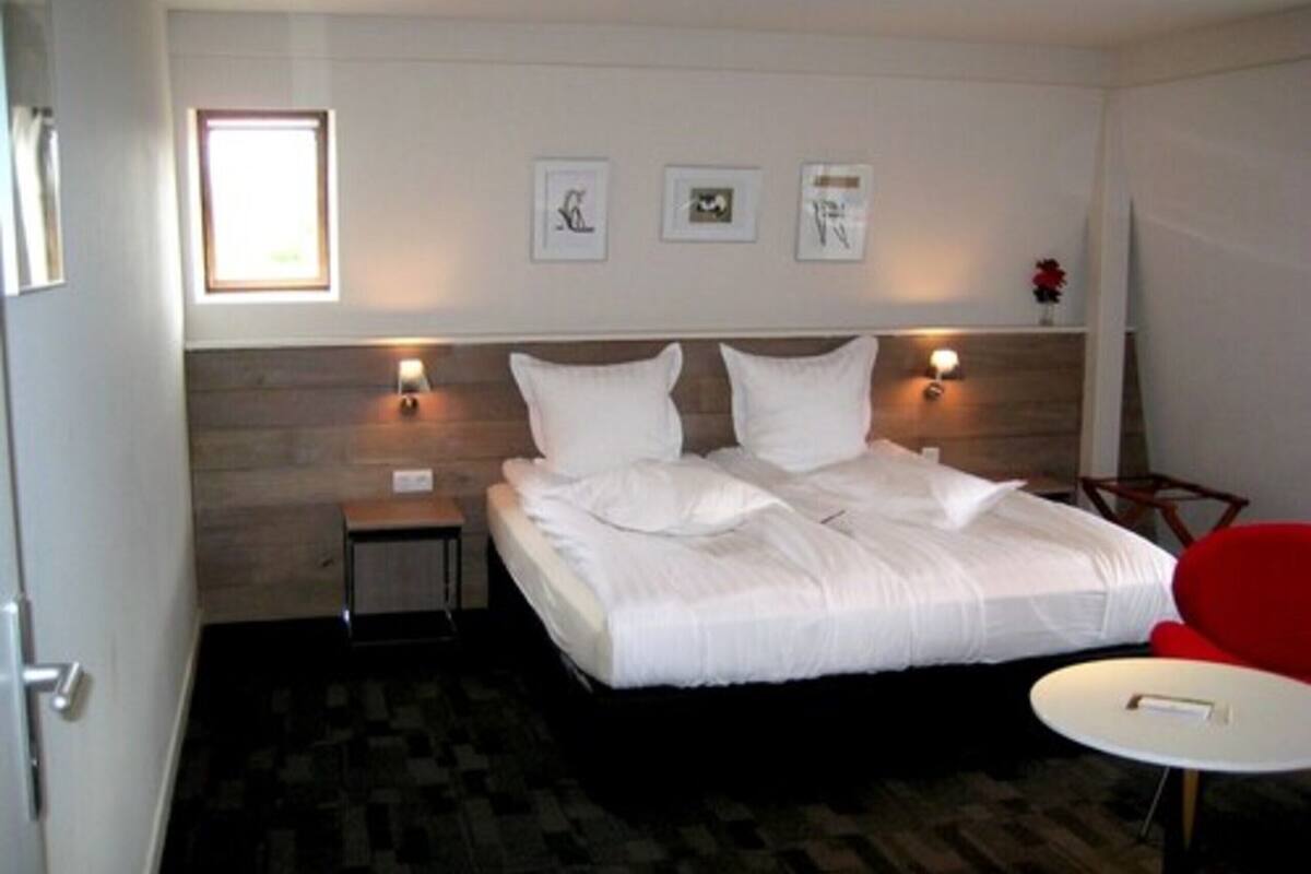 A hotel room with a bed, a small bedside table, a chair, and a round table. There are wall-mounted lights and framed pictures on the wall.