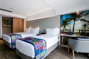 A hotel room with two beds, a desk, a chair, a painting of a beach scene, and a small table with a bottle and a phone. - Arena Ipanema Hotel (Rio de Janeiro)
