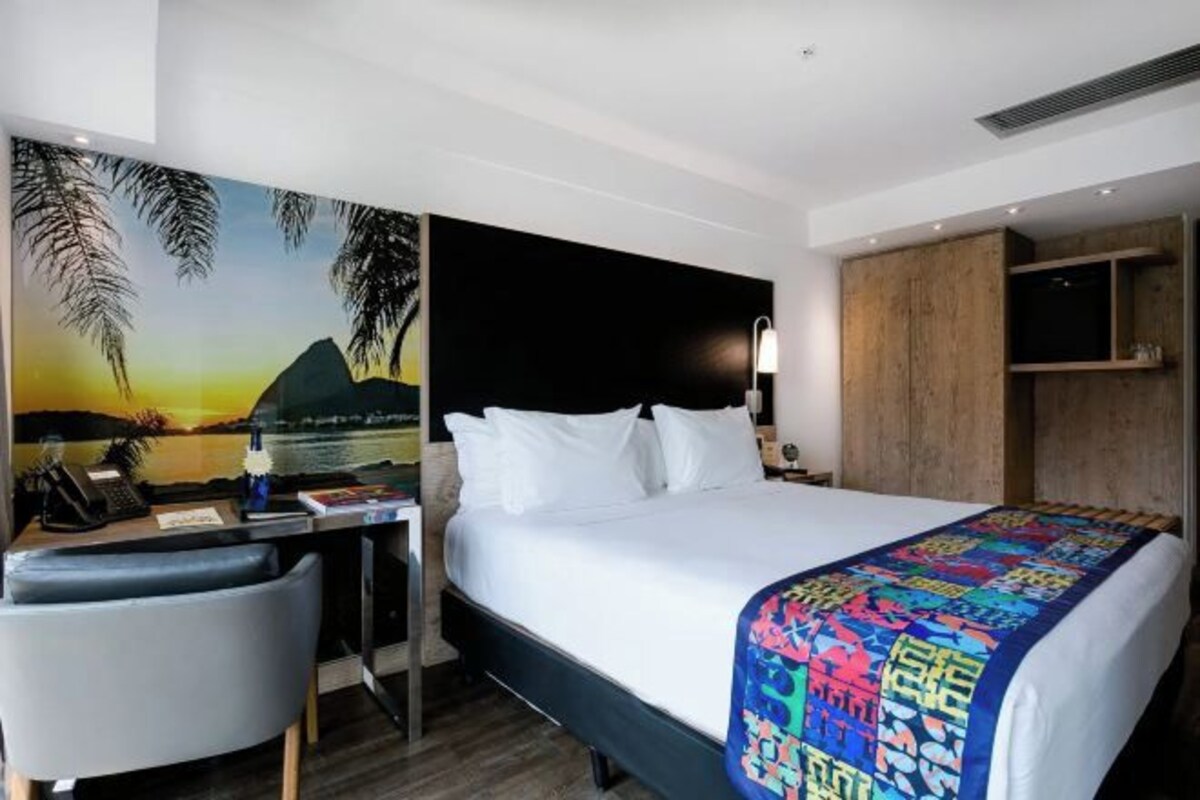 A hotel room with a bed, desk, chair, and a painting of a sunset.
