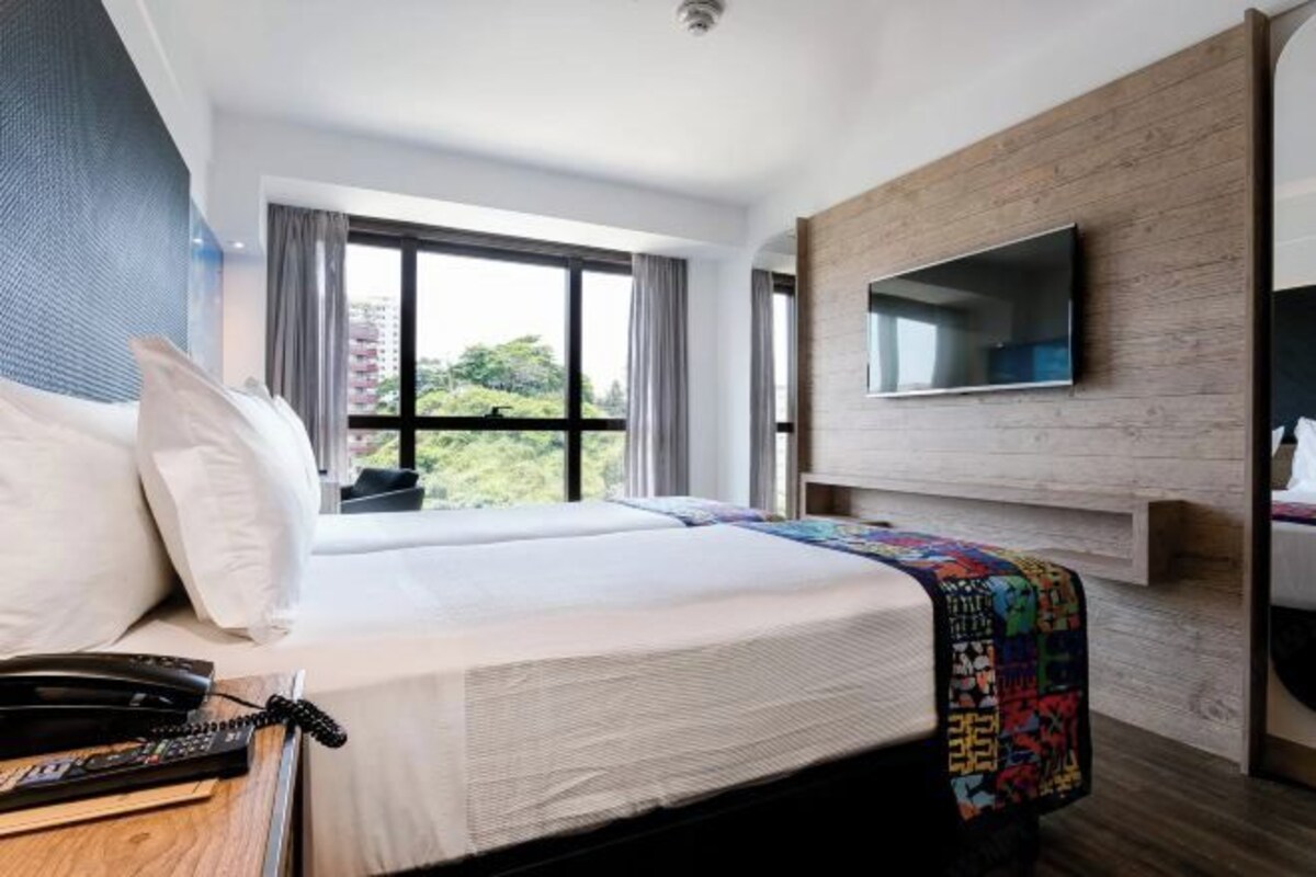 A hotel room with a large bed, a flat-screen TV mounted on the wall, a bedside table with a telephone, and a window offering a view of greenery.