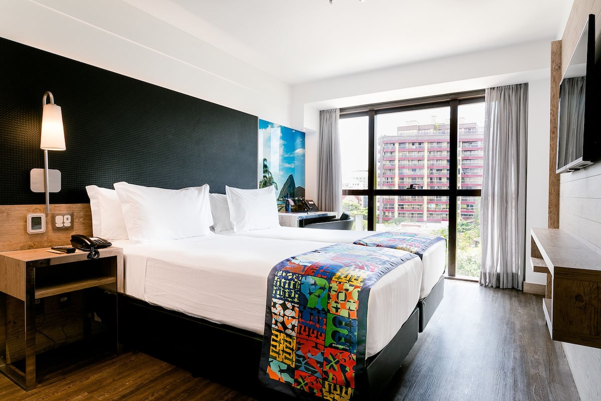 Una habitación de hotel con cama, mesita de noche, lámpara y un ventanal con cortinas.