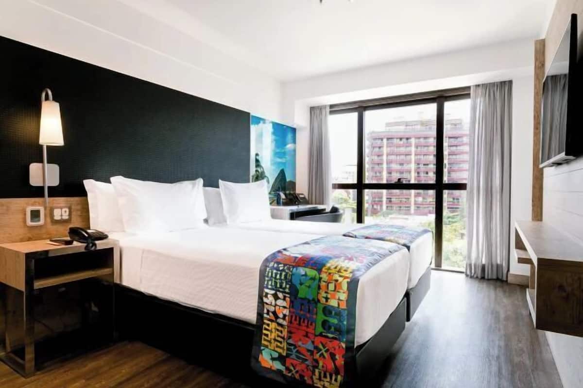 Habitación de hotel con cama, mesita de noche, lámpara y un amplio ventanal con cortinas.