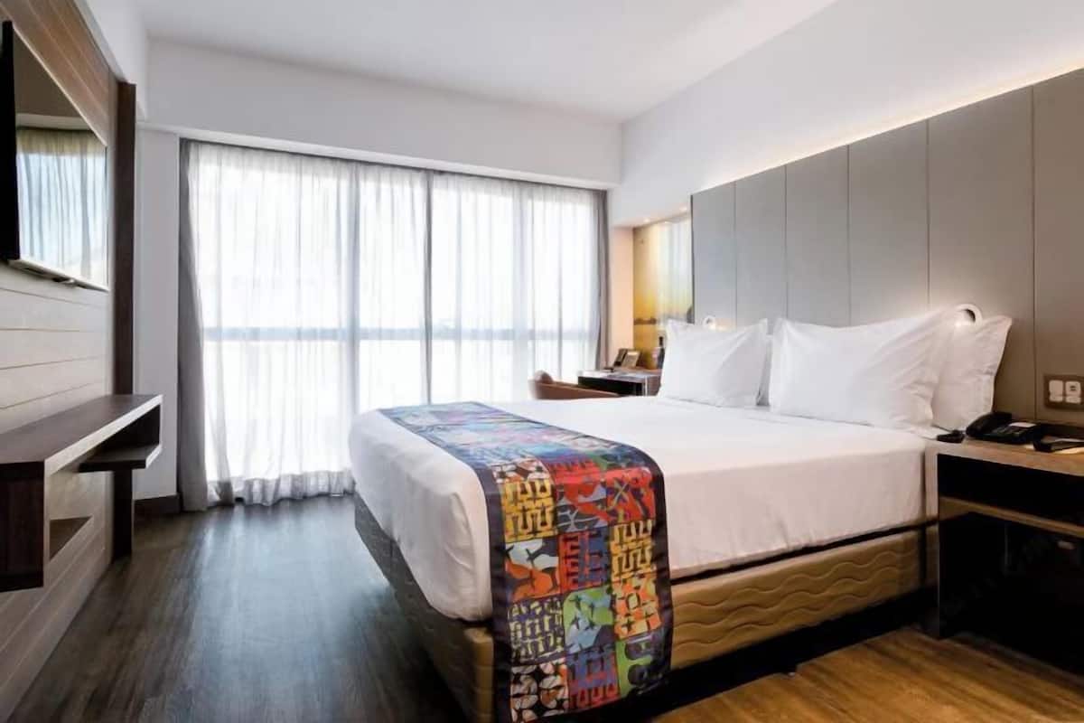 Habitación de hotel con cama, escritorio, silla, televisor y ventana con cortinas.