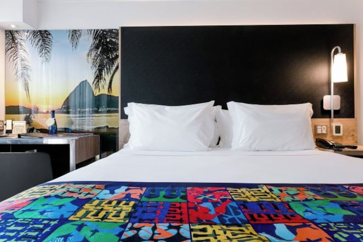 A hotel room with a bed, a desk, a nightstand, a lamp, and a wall with a tropical mural.