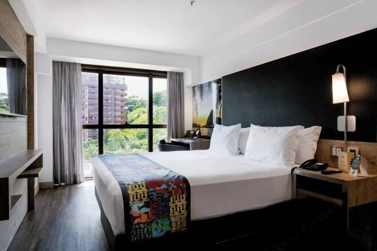 Una habitación de hotel con una cama grande, un escritorio, una silla, una mesita de noche, una lámpara y una ventana con cortinas.